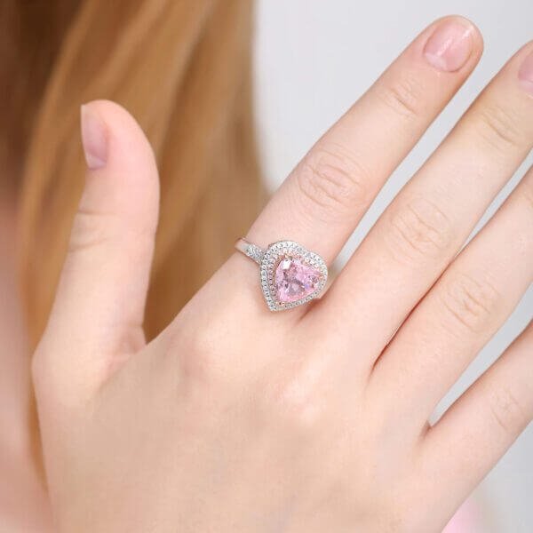 A hand adorned with a Romantic Heart Halo Ring, showcasing a pink gemstone encircled by tiny clear stones on a sleek silver band. The neutral background highlights the intricate design, drawing attention to its enchanting elegance.