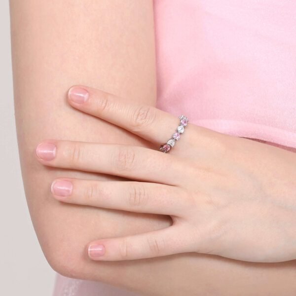 A person wearing a pink top is proudly displaying an Eternal Love Ring adorned with multicolored gemstones on their finger, crossing their arm over their chest. Their nails are neatly manicured.