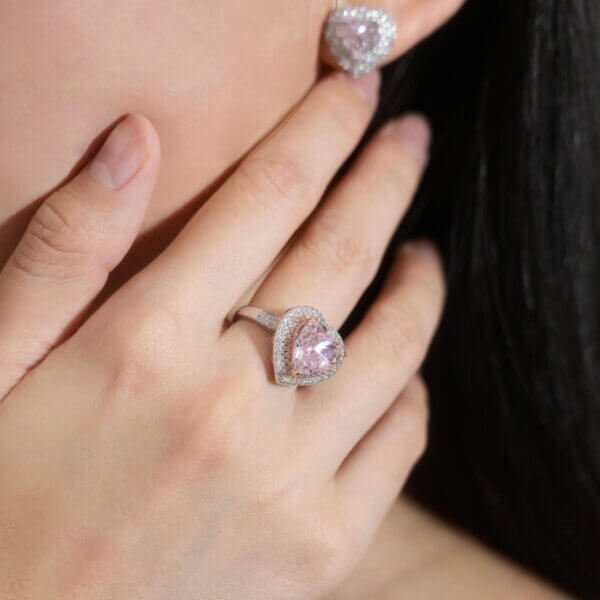 A person wearing a romantic heart halo ring with a heart-shaped pink gemstone, framed by small diamonds, on their ring finger. Matching heart-shaped pink earrings complete the look as their hand gently touches their face.