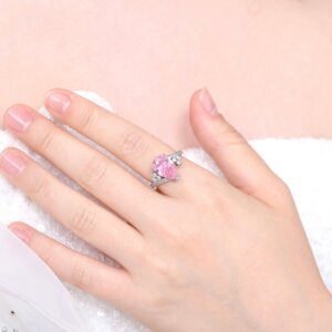 A hand showcases a stunning Pink Passion Pear Ring, featuring a heart-shaped pink gemstone, accented with smaller clear stones on the band. The silver ring rests elegantly on a white, textured surface, possibly a piece of clothing.