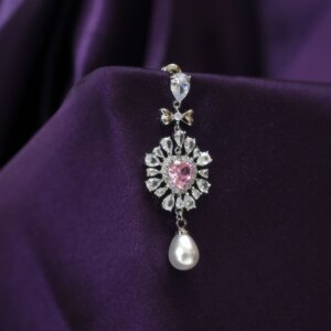 A luxurious pendant against a deep purple fabric background. The pendant features a pink heart-shaped gem surrounded by clear diamonds in a sunburst pattern, with a teardrop pearl dangling at the bottom.