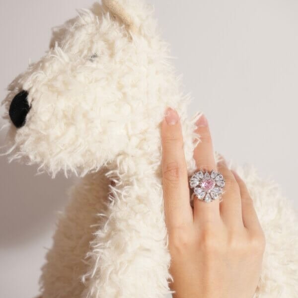 A hand with a large, ornate ring featuring a pink center stone and surrounding clear stones is resting on a fluffy white teddy bear. The background is a plain light color.