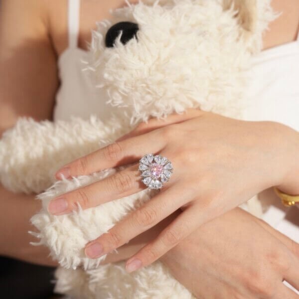 A person with light skin holds a fluffy white teddy bear. On their hand, they wear a large, intricate ring with a floral design, featuring sparkling gems. The overall setting is soft and warm, highlighting the jewelry.