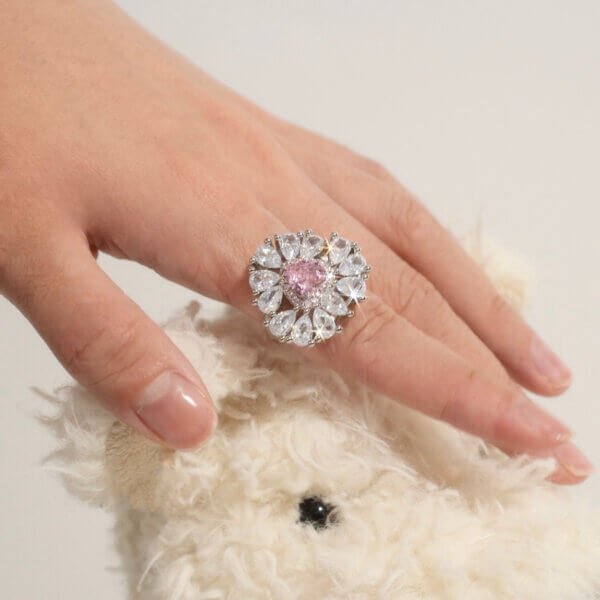 A hand wearing a large, ornate ring with clear and pink stones is gently resting on a fuzzy white teddy bears head. The rings design resembles a flower, and the setting has a luxurious appearance.