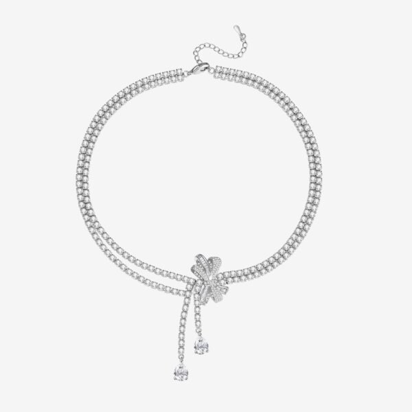 A sparkling diamond necklace with two strands converging at a floral-shaped cluster. The cluster features a bow-like design accented by two dangling pear-shaped diamonds. The background is a plain, light color.