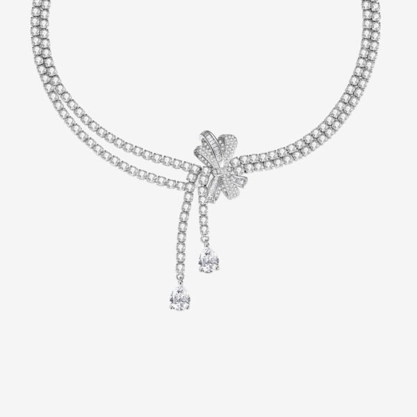 A sparkling diamond necklace with two strands converging at a floral-shaped cluster. The cluster features a bow-like design accented by two dangling pear-shaped diamonds. The background is a plain, light color.