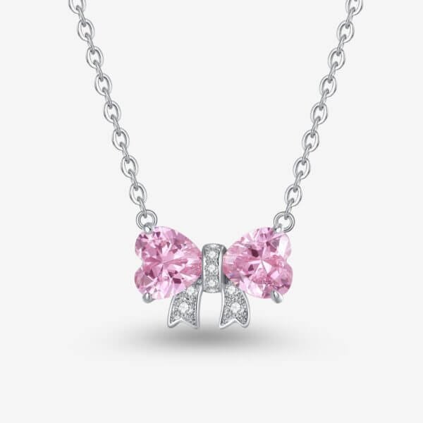 A silver necklace with a bow-shaped pendant. The bow features two pink heart-shaped gemstones as loops and small white stones lining the knot, against a plain white background.