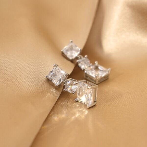 Close-up of silver square-shaped earrings with clear gemstones, resting on a smooth, satin gold fabric.