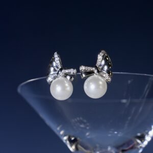 Elegant pearl earrings with bow-shaped, diamond-studded settings, displayed on the rim of a clear glass against a dark blue background.