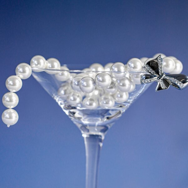 A string of white pearls drapes over a clear martini glass filled with more pearls, against a blue background. A decorative silver bow is attached to the string, enhancing the elegant and luxurious aesthetic.
