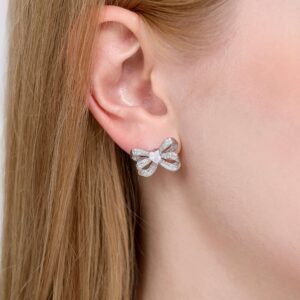 Close-up of a person with straight blonde hair wearing a sparkling, diamond-encrusted bow-shaped earring on their left ear. The earring reflects light, highlighting its intricate design.