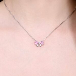 A close-up of a silver necklace with a delicate pink bow pendant adorned with small gemstones, worn around a persons neck. The background is a soft, neutral skin tone.