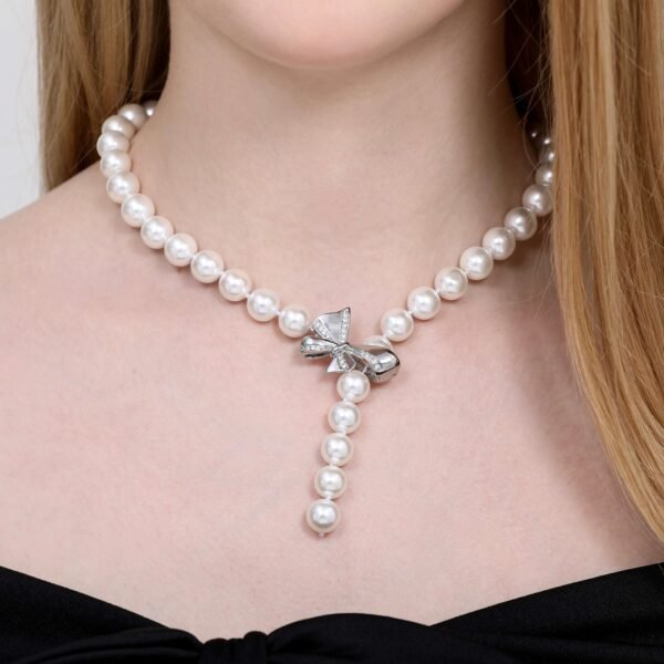 A person wears a pearl necklace with a silver bow-shaped clasp, creating a Y-shape. The pearls are round and evenly spaced, and the person is dressed in a black, off-the-shoulder top. The background is neutral.