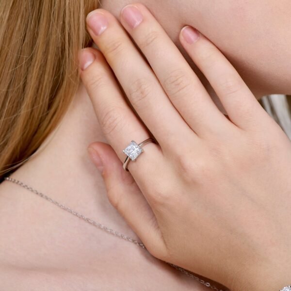 A close-up of a person wearing a silver ring with a large, square-cut diamond on their ring finger. Their hand is gently touching the side of their face, and they are wearing a delicate silver necklace.