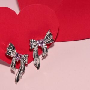 Silver earrings shaped like bows, adorned with sparkling crystals and featuring a pink heart-shaped gem in the center. The design is delicate and elegant.